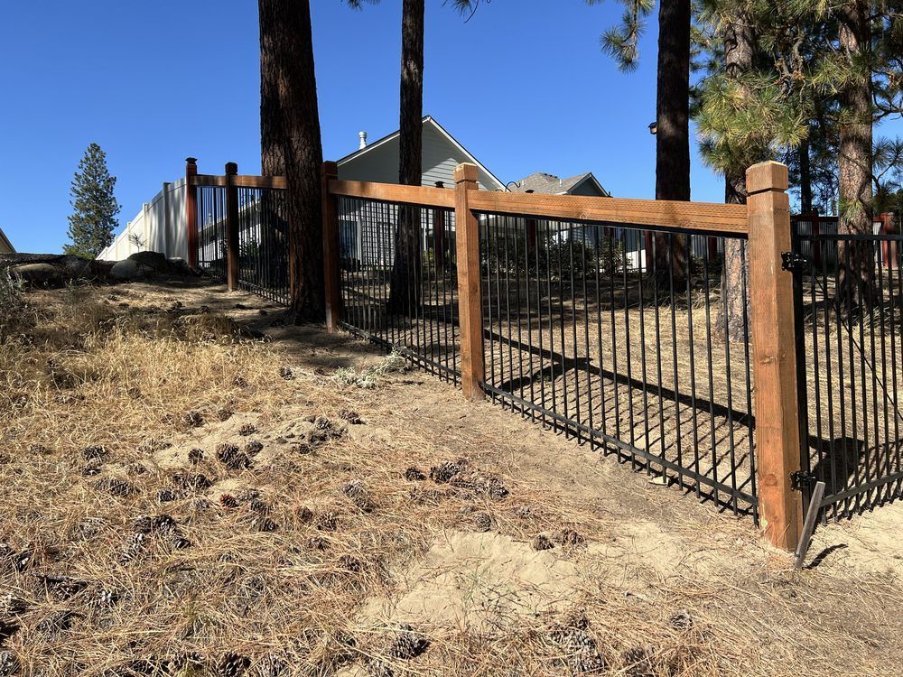 Black metal gate and fence beside a dry, wooded yard with a house in the background.