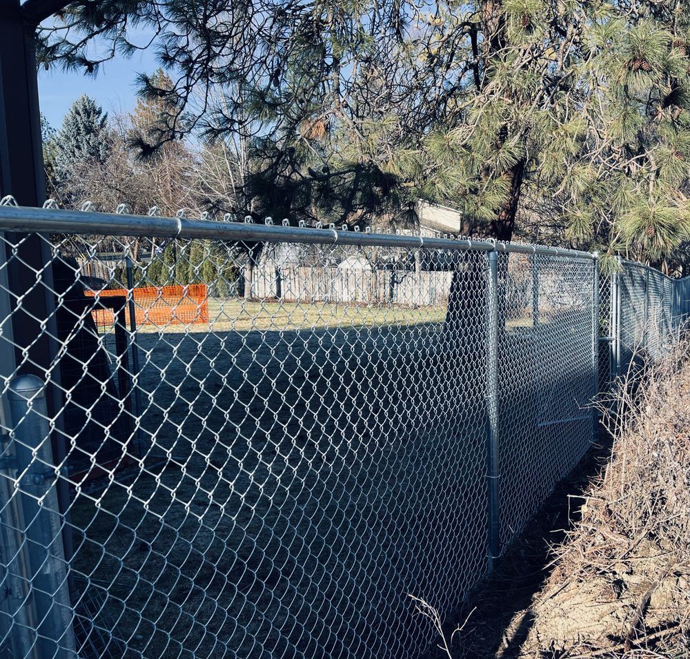 Chain-link fence beside a wooded area, with a trash bin and orange barrier in the background