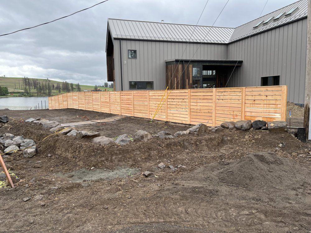 Gray industrial building with a wooden fence around a gravel and dirt construction area under a cloudy sky