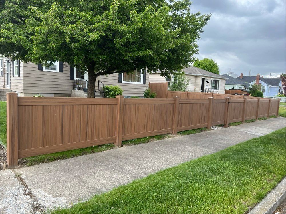 Brown wooden fence along a concrete sidewalk beside a house under cloudy skies.