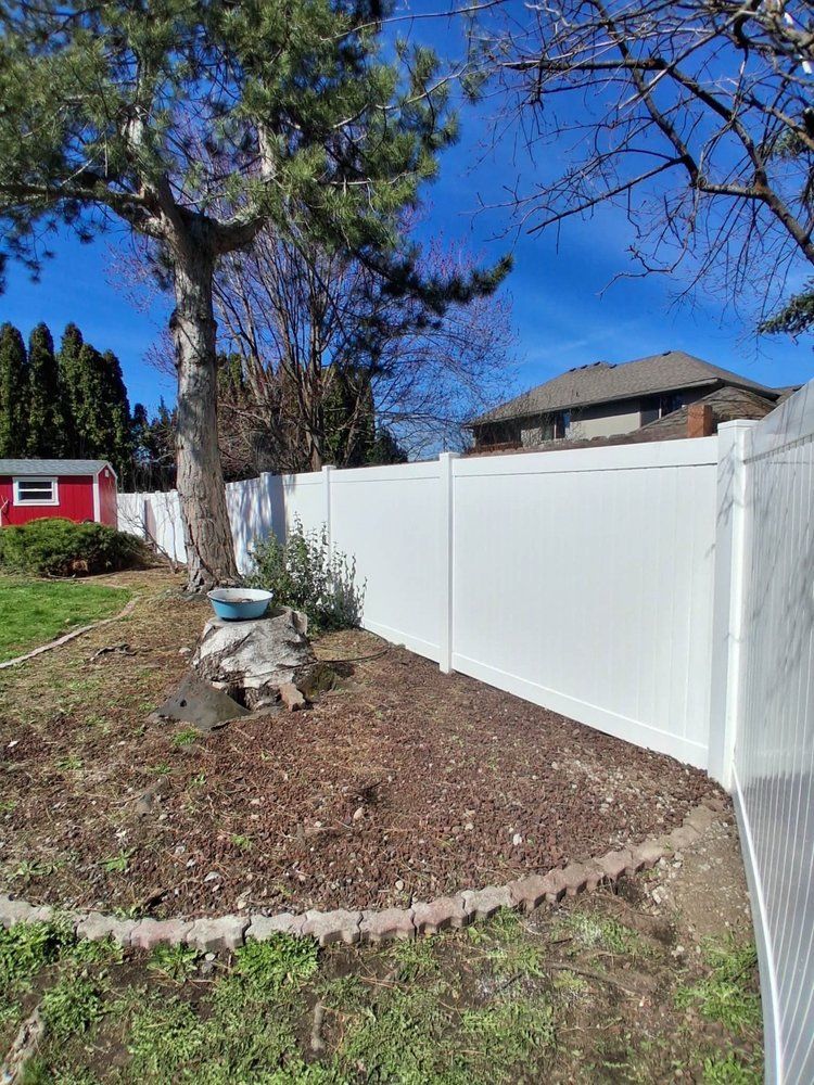 White vinyl fence along a grassy yard with a tree stump, trees, and a red shed under a clear blue sky
