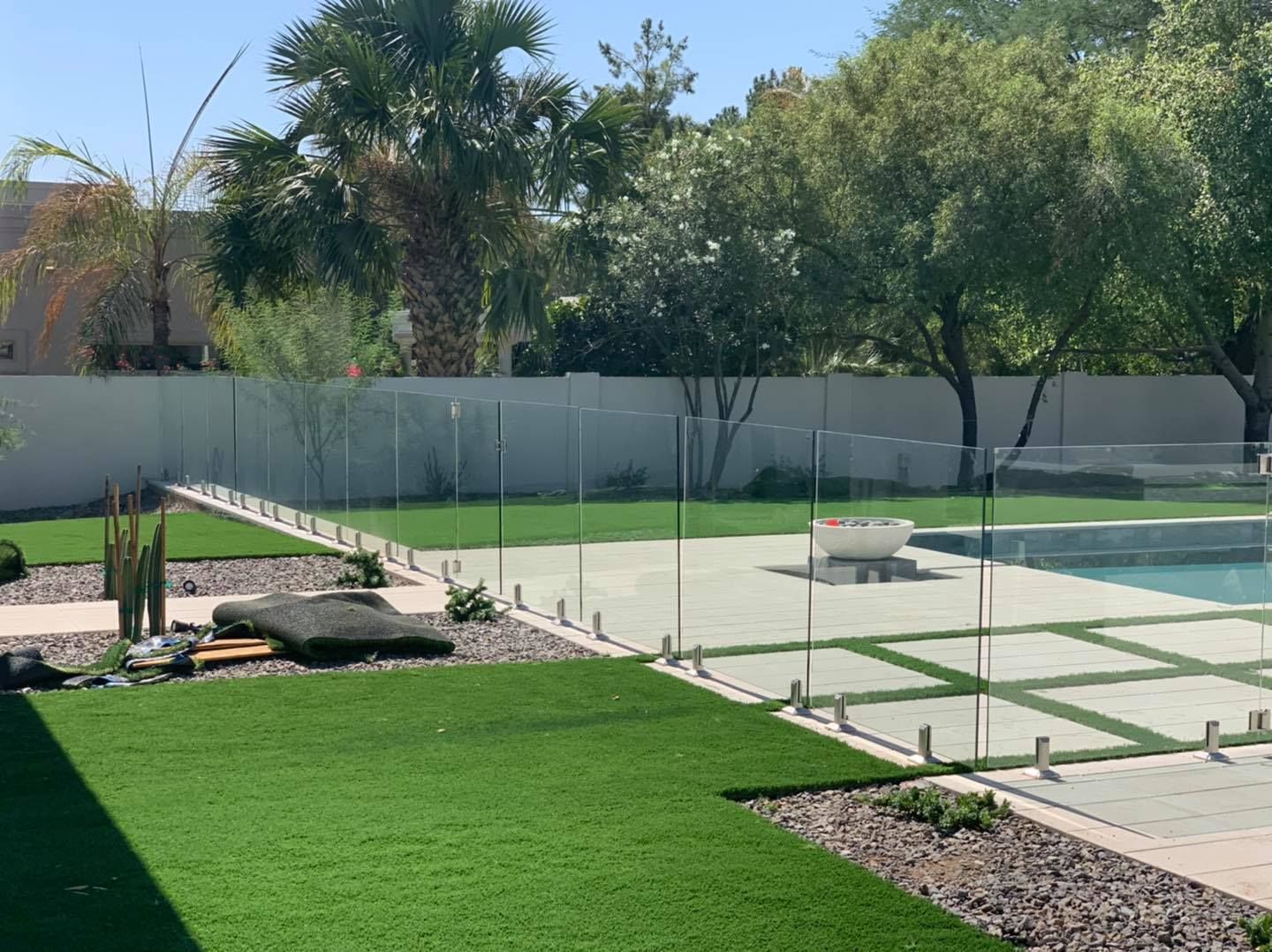 Glass fence surrounds a backyard with pool, artificial turf, and landscaping under a sunny sky.
