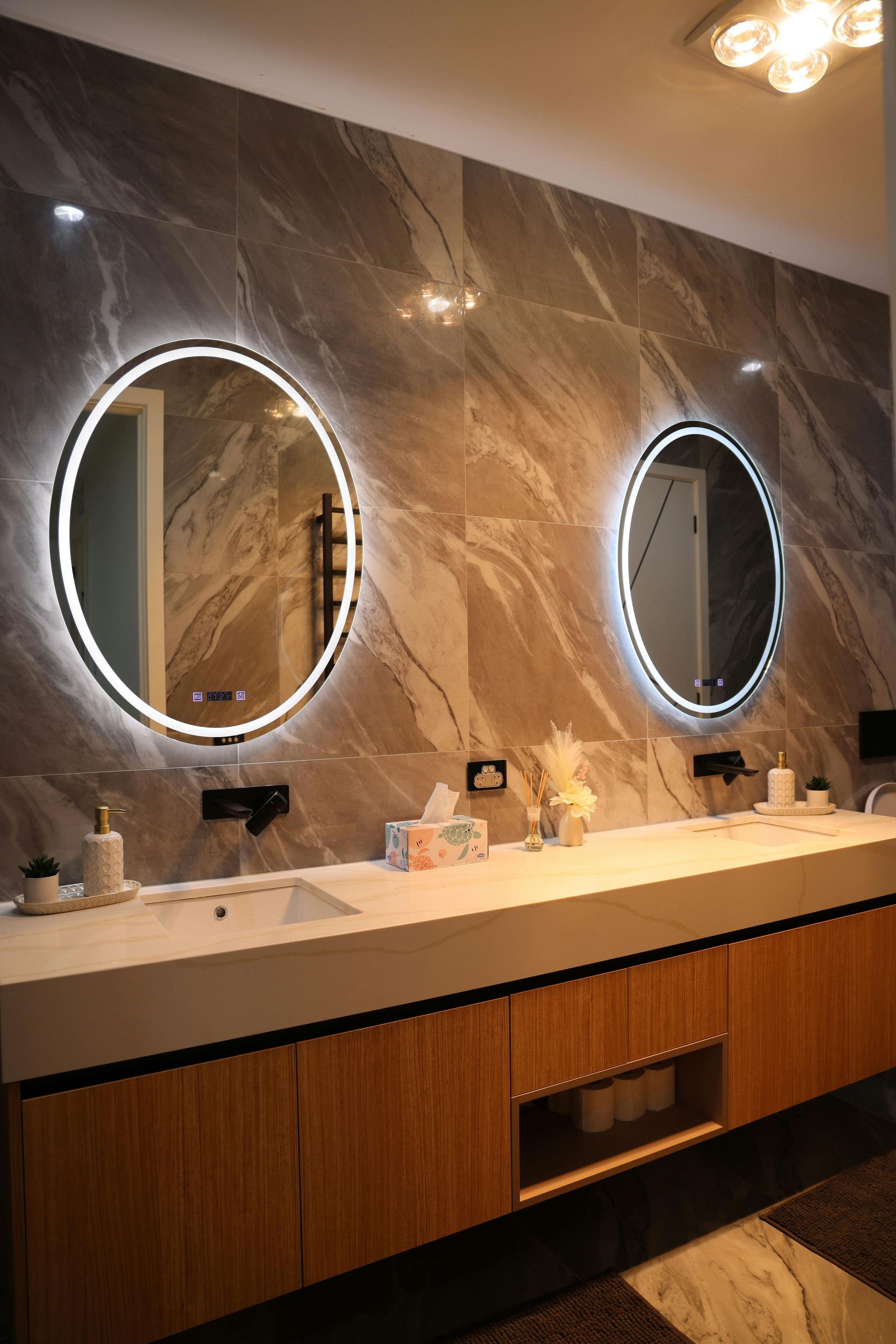 A modern bathroom vanity featuring a long white counter, dual backlit circular mirrors, and wooden cabinetry.