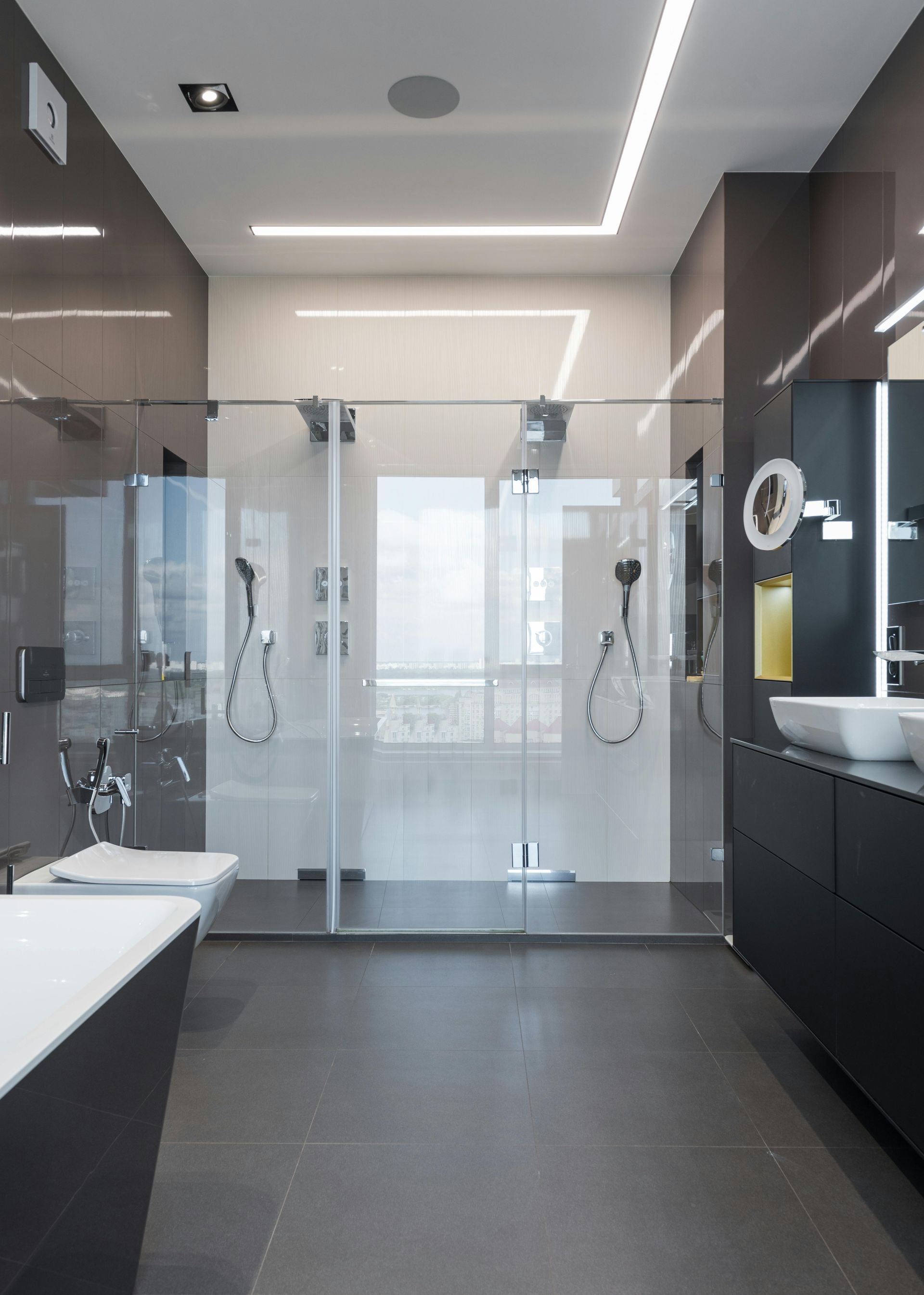 Modern bathroom with a glass-enclosed double shower, dark grey cabinetry, white sinks, and recessed ceiling lighting.