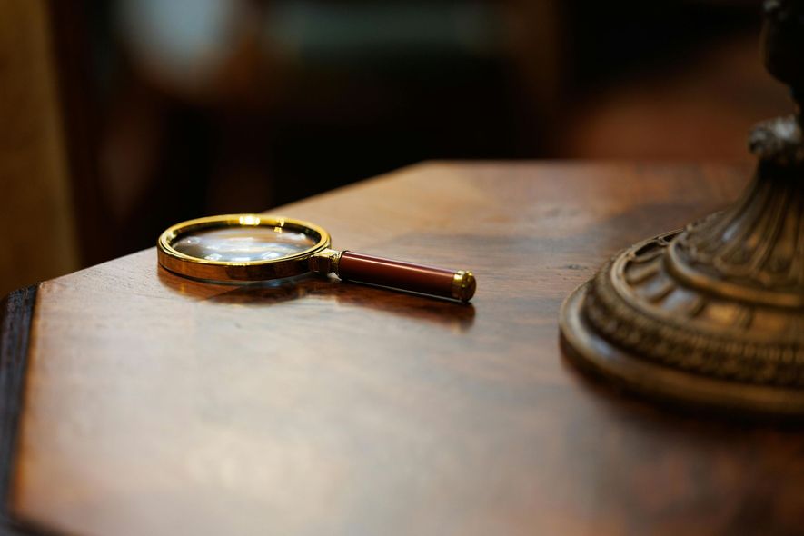 A magnifying glass rests on a polished wooden surface next to the base of an antique lamp.