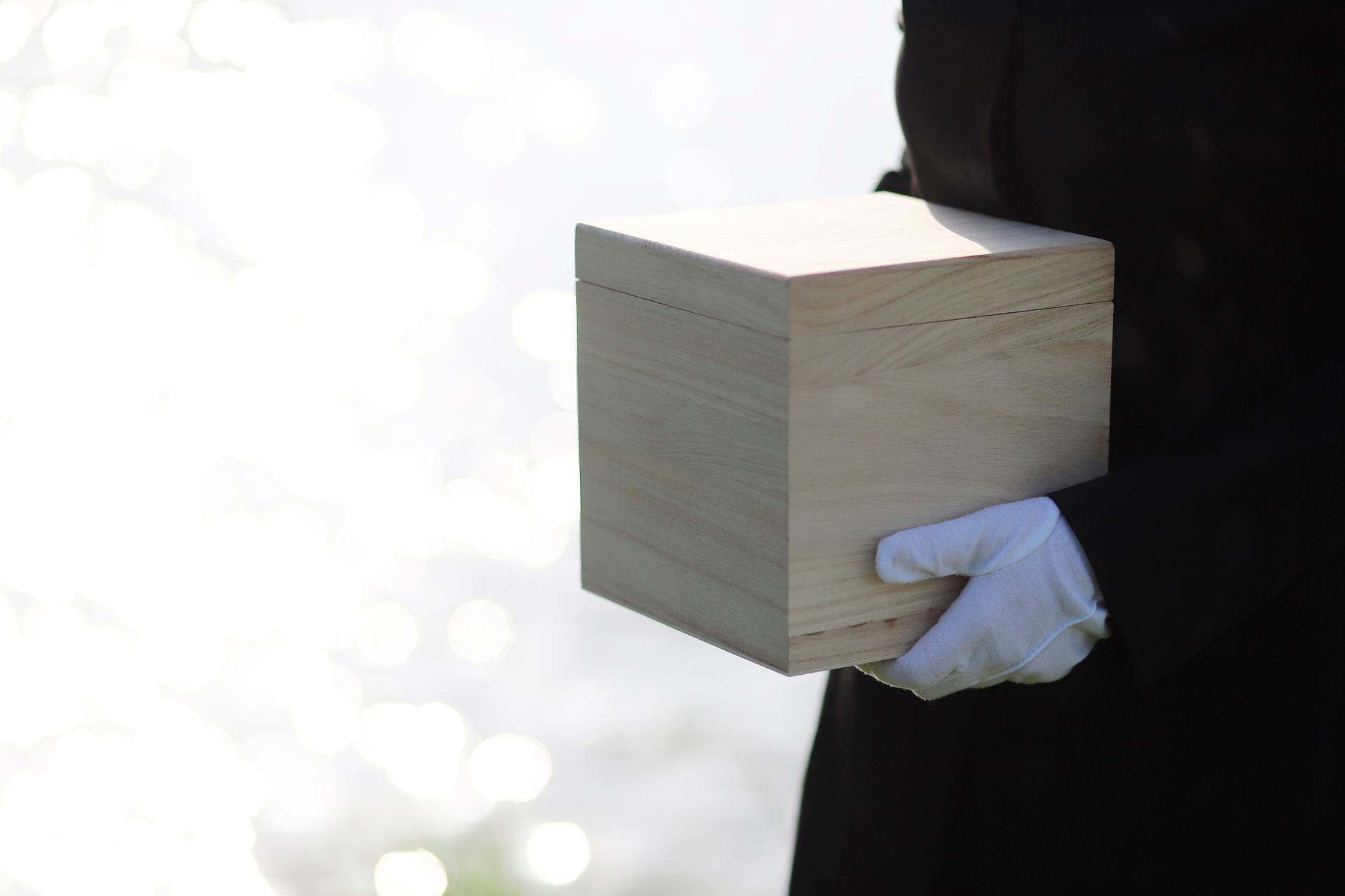 Person in black suit and white gloves holding a light wooden cremation urn outdoors near water.
