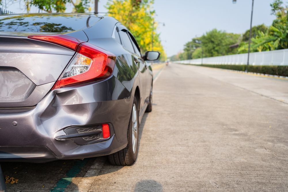 A grey car parked on a roadside with a noticeable dent on its rear bumper.