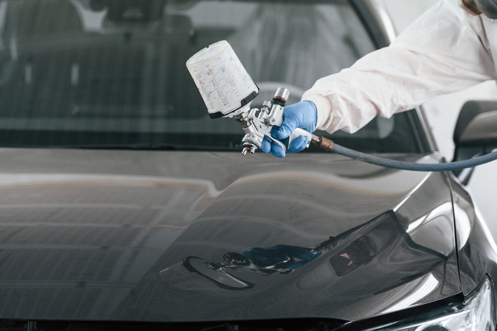 A worker in protective gear uses a paint spray gun to apply a finish to the hood of a dark-colored vehicle.
