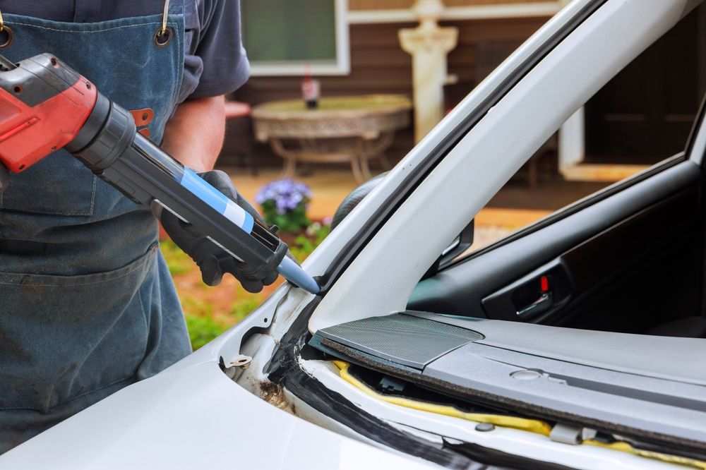 A worker in a denim apron uses a red caulking gun to apply adhesive to a car windshield frame.