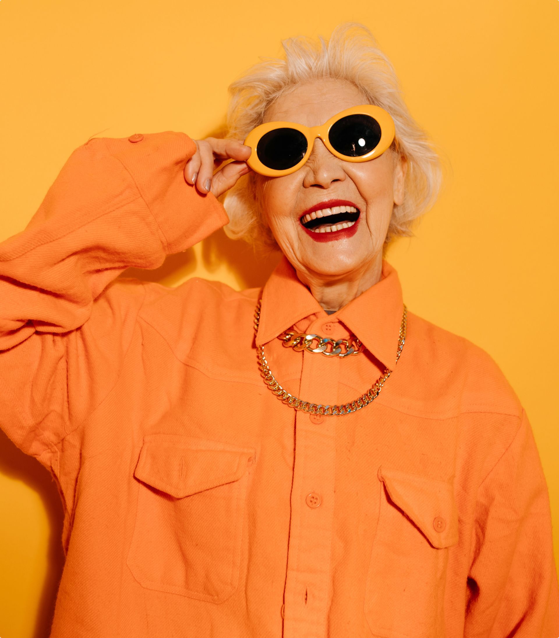 Elderly woman with white hair, wearing orange shirt and sunglasses, smiling in front of a yellow background.