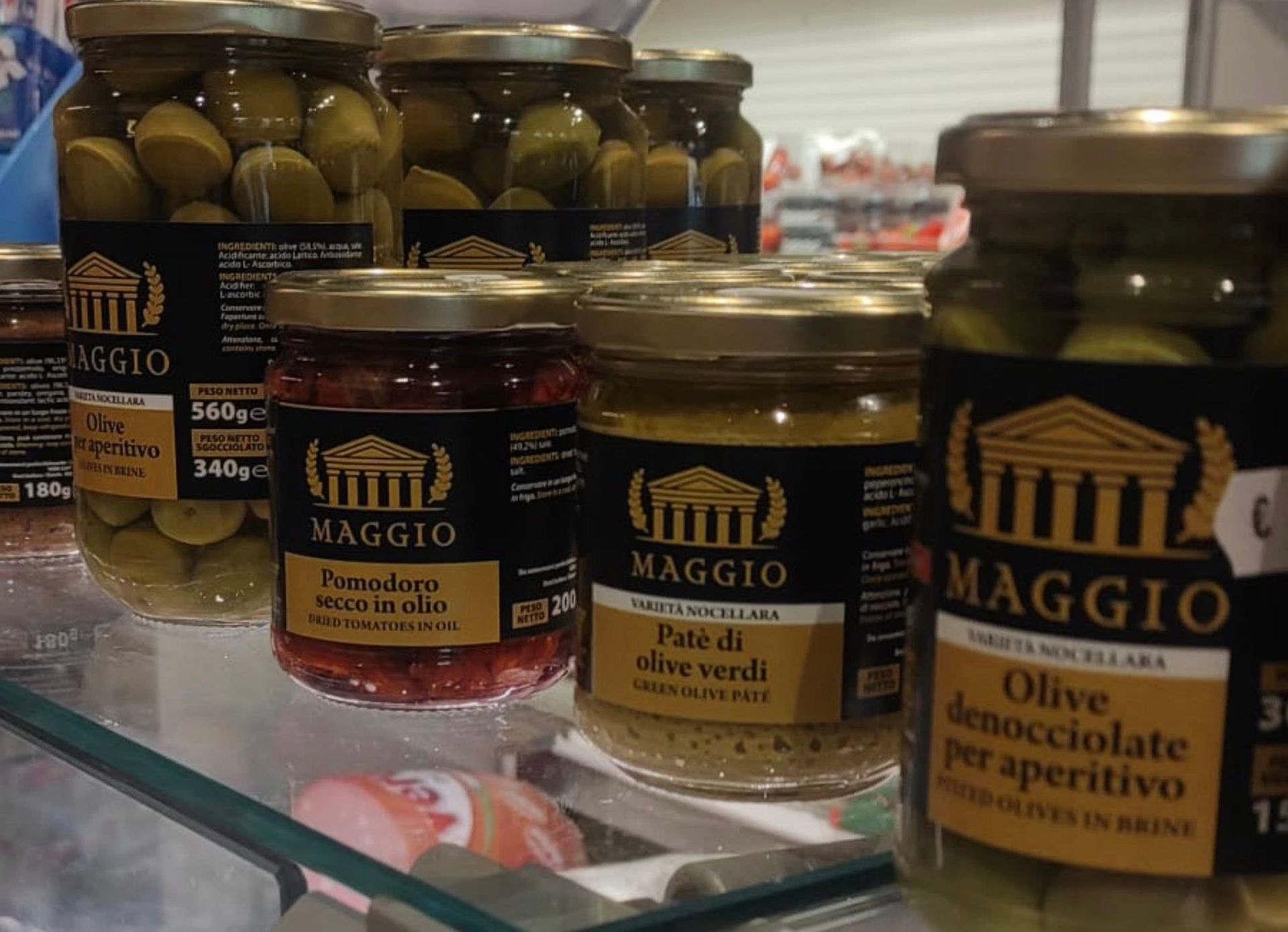 Su uno scaffale di un negozio, vasetti di vetro contenenti olive Haggio, tra cui olive verdi e peperoncini in salamoia rossa.
