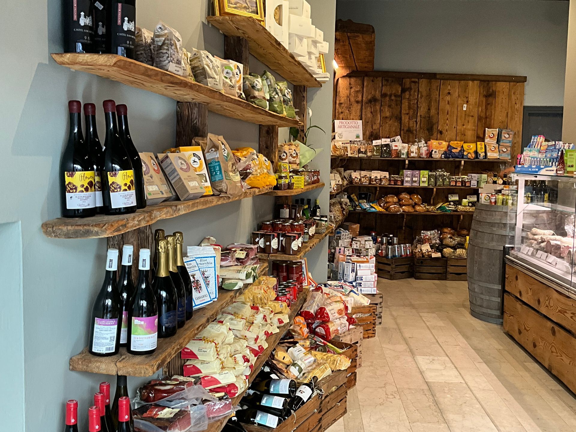 Piccolo corridoio di generi alimentari con bottiglie di vino, snack confezionati e scaffali in un interno di negozio rustico
