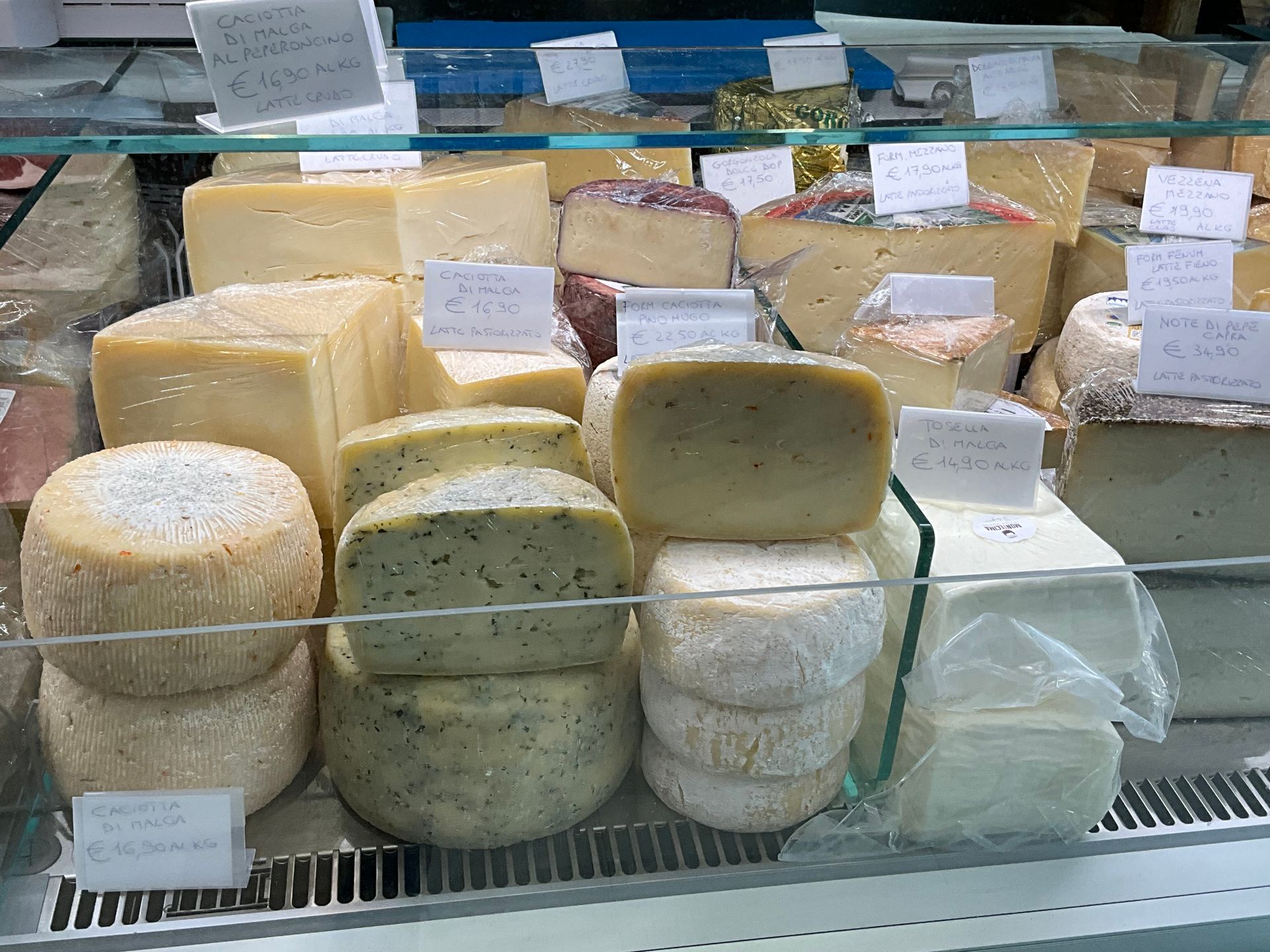 Forme e blocchi di formaggio esposti in una vetrina refrigerata con etichette dei prezzi.