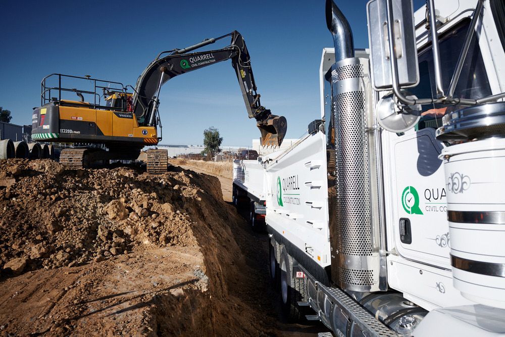 A yellow excavator loads dirt into the white dump truck at an outdoor construction site under a clear blue sky Quarrell Civil Construction In Congupna, VIC