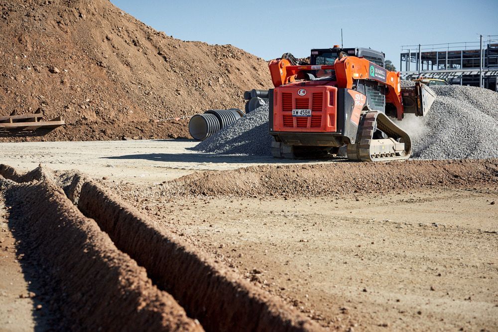 An Orange Tracked Compact Loader Moves a Pile of Gravel on a Sunny Construction Site — Quarrell Civil Construction In Congupna, VIC