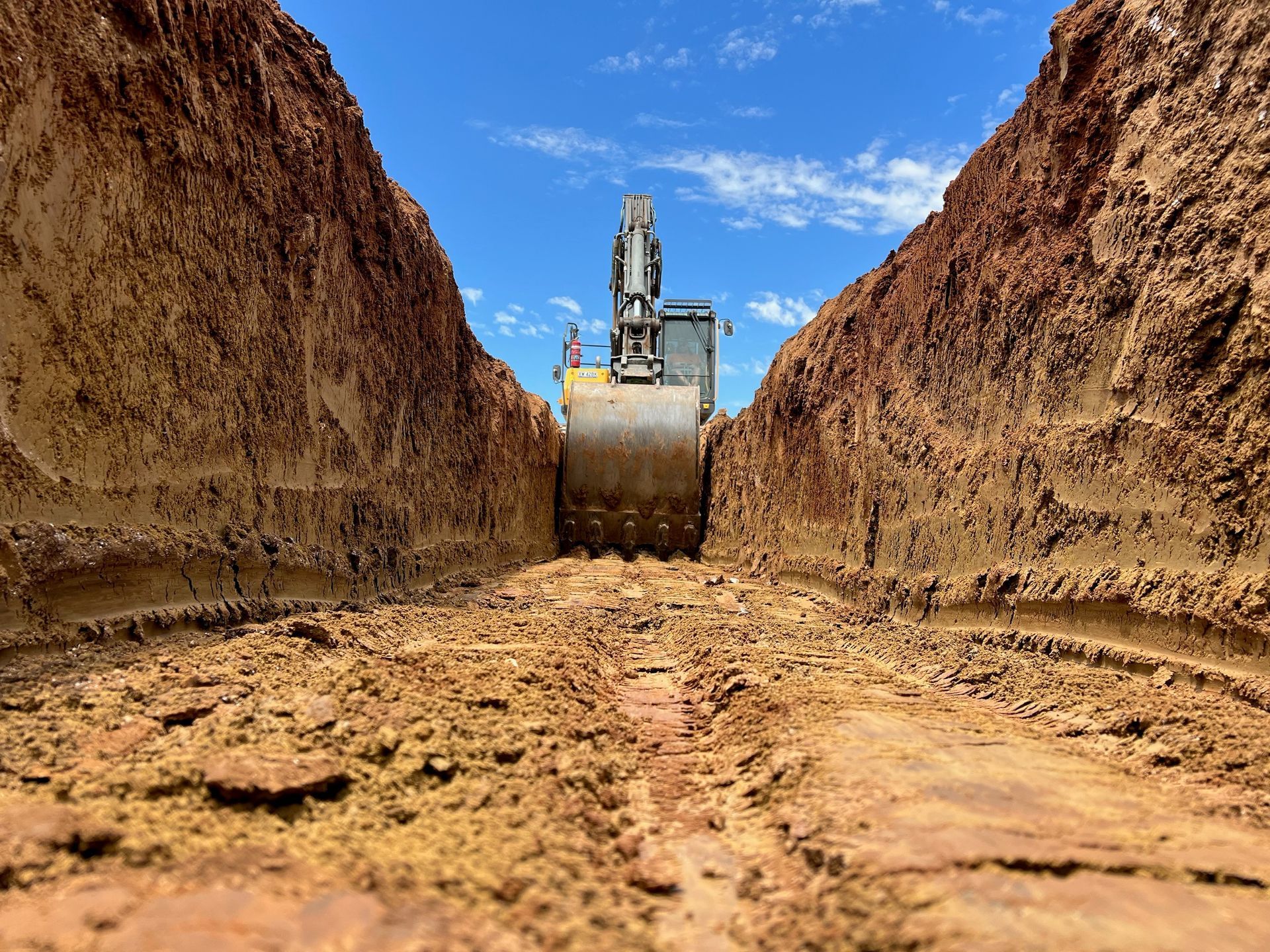 An excavator digging a deep, narrow trench in reddish-brown soil under a bright blue sky  — Quarrell Civil Construction In Congupna, VIC
