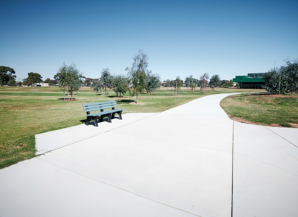 Park with a light blue bench, concrete path, green grass, trees, and a building on a sunny day. — Quarrell Civil Construction In Congupna, VIC