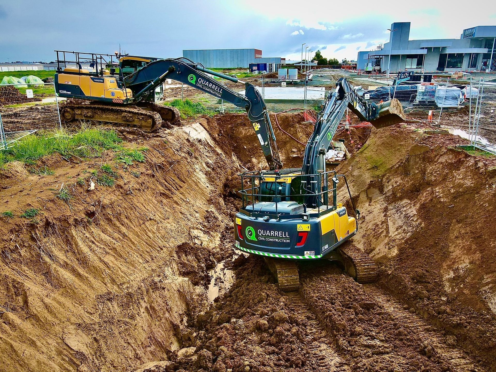 Two yellow and grey excavators work at the bottom of a deep muddy construction trench under an overcast sky — Quarrell Civil Construction In Congupna, VIC