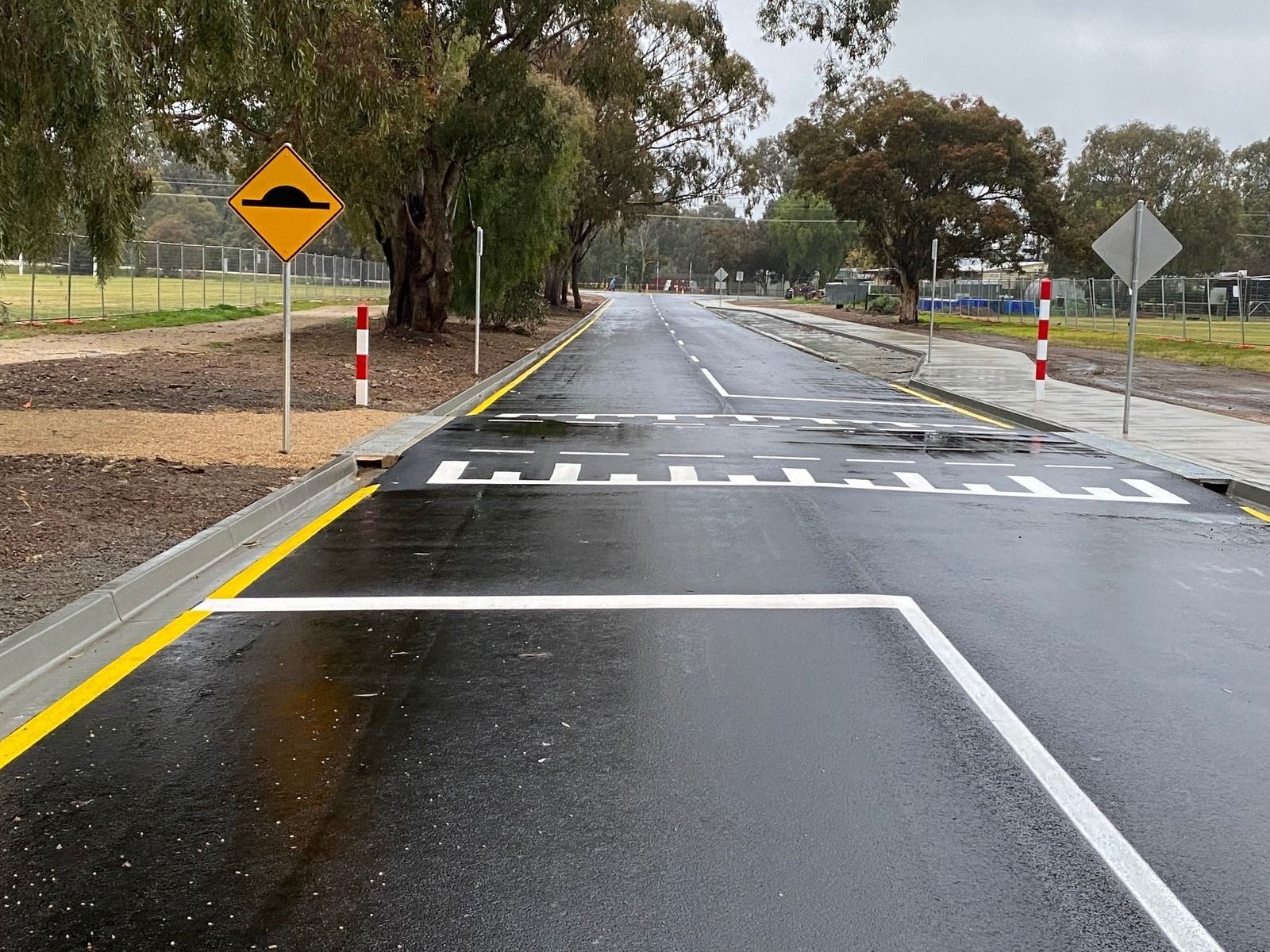 A wet, paved road with a speed bump marked by white lines, a yellow warning sign, and orange-and-white bollards - Quarrell Civil Construction In Congupna, VIC