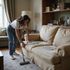 Woman vacuuming a beige couch in a living room; she wears a white shirt and jeans.