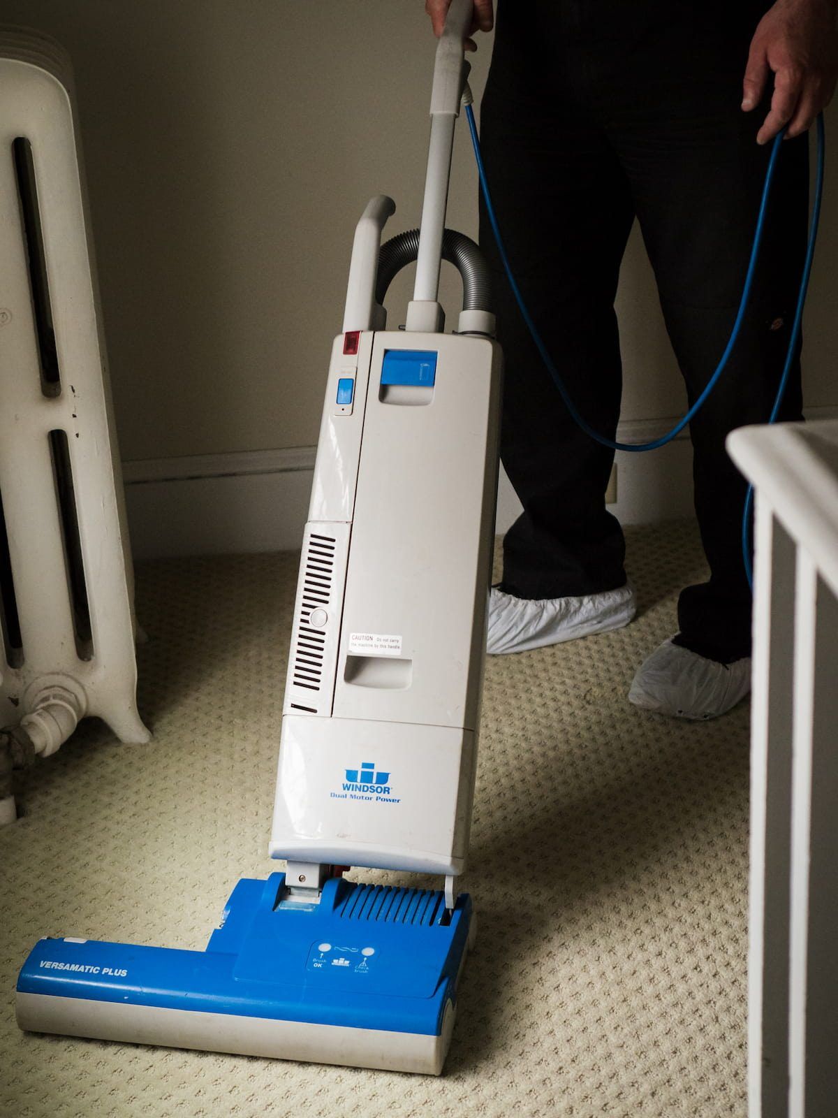 Ray DeMeo, wearing shoe covers, vacuuming a carpeted room in a home.