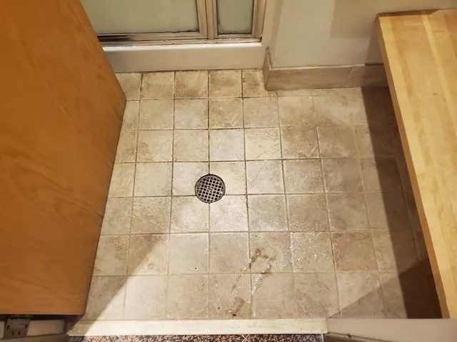 Textured tile floor with center drain outside an office building shower, with soil in the tile texture and recessed grout lines before cleaning