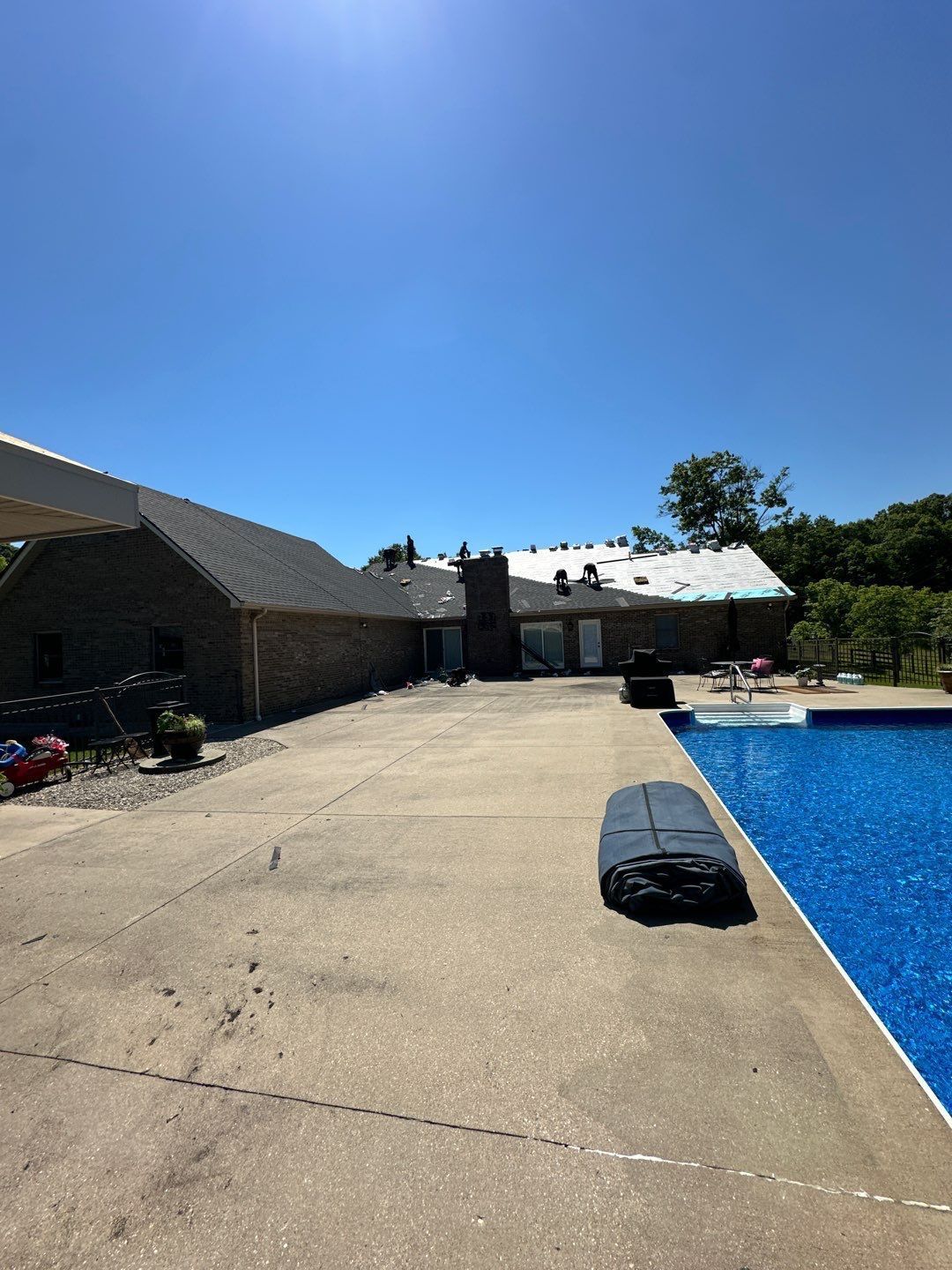 Sunny backyard patio with blue swimming pool, concrete deck, lounge chairs, and brick house.