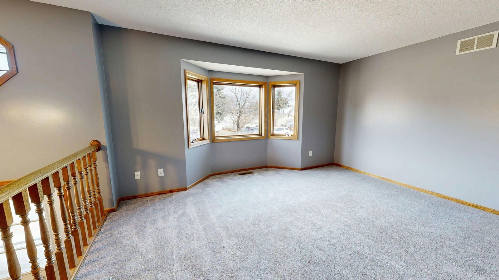 Empty carpeted living room with gray walls, bay window, and wooden stair railing.