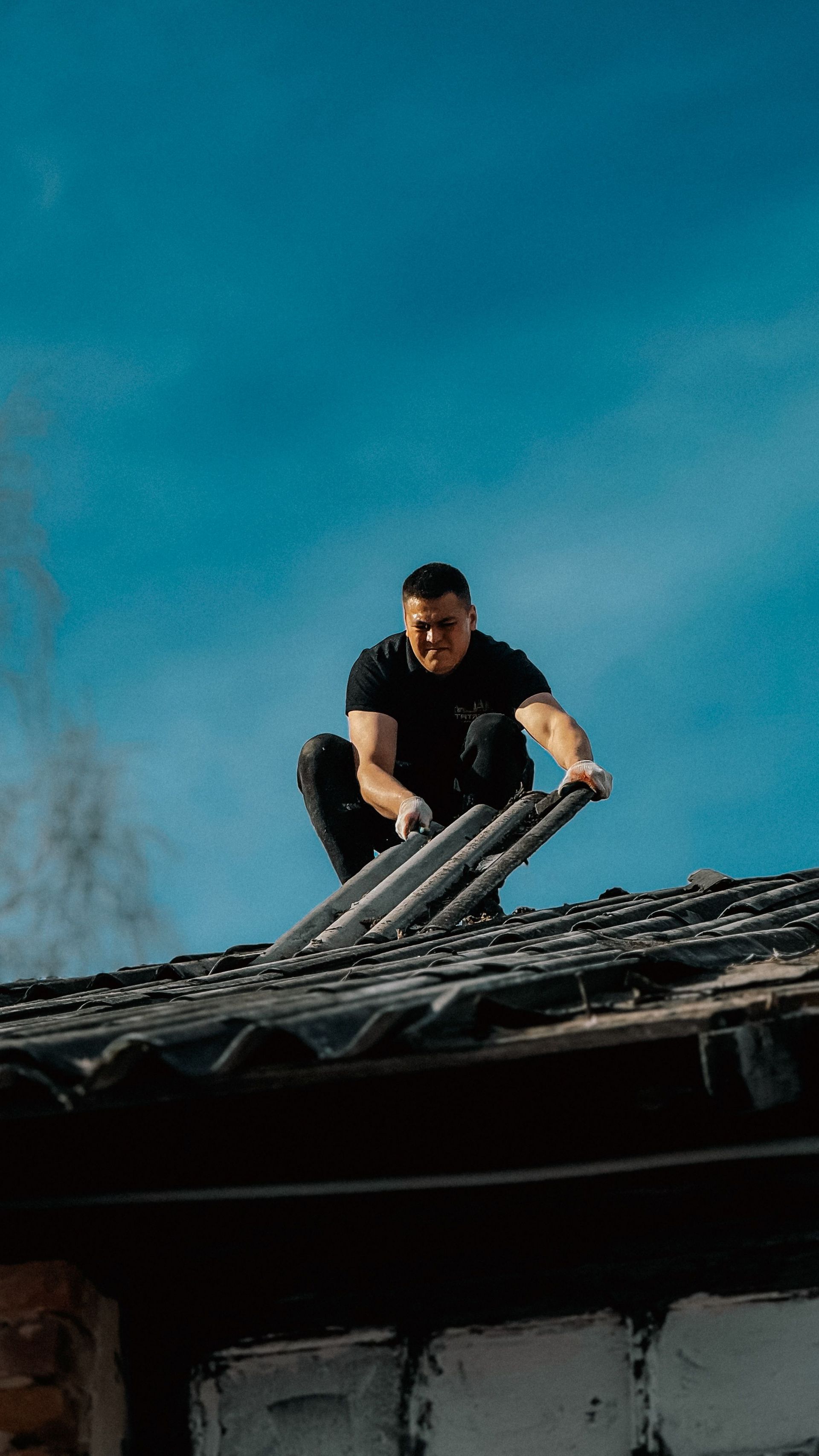 Worker on a roof installing dark shingles against a bright blue sky.