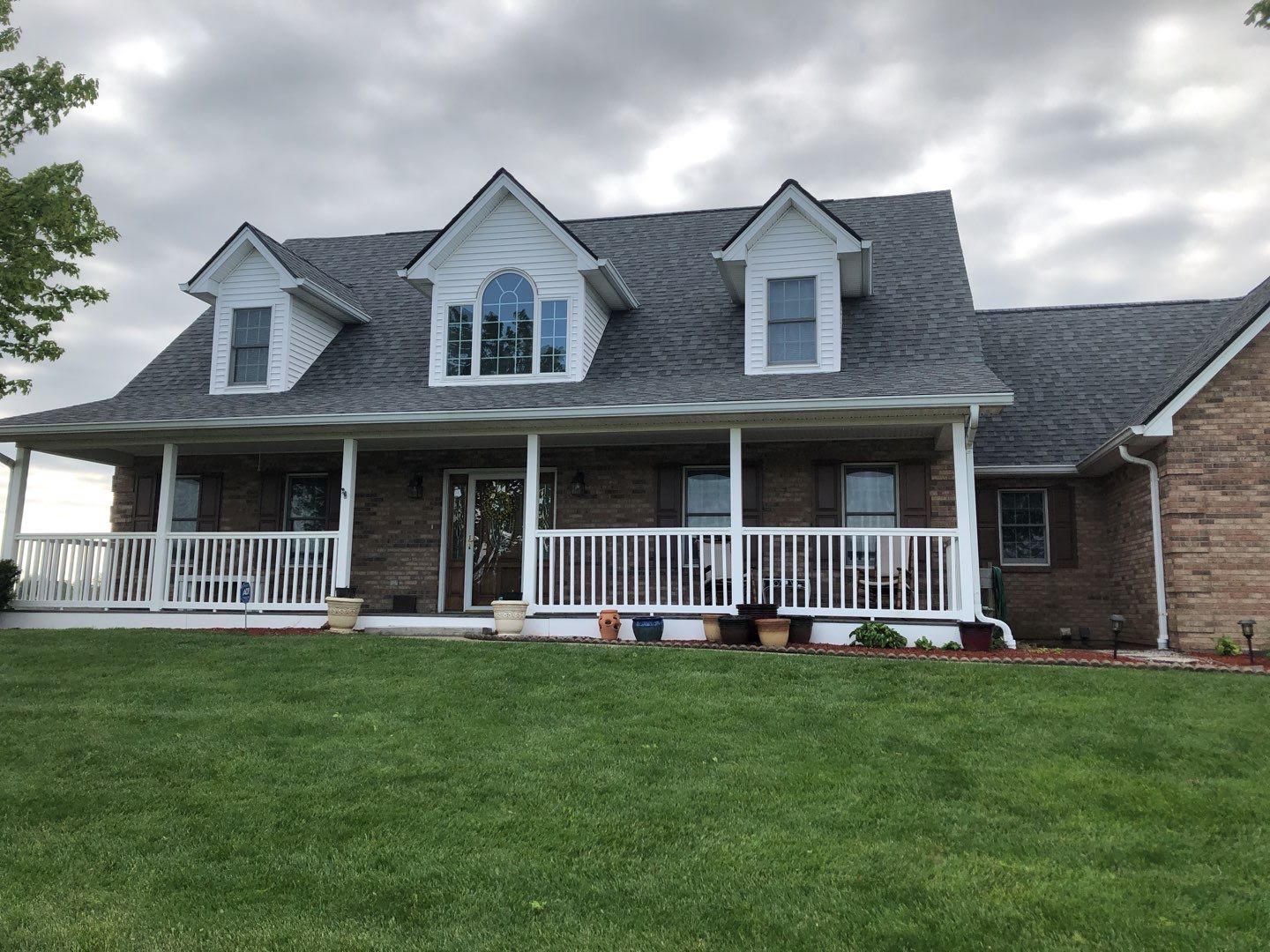 Two-story suburban house with white porch railings, dormer windows, and a large green lawn under cloudy skies