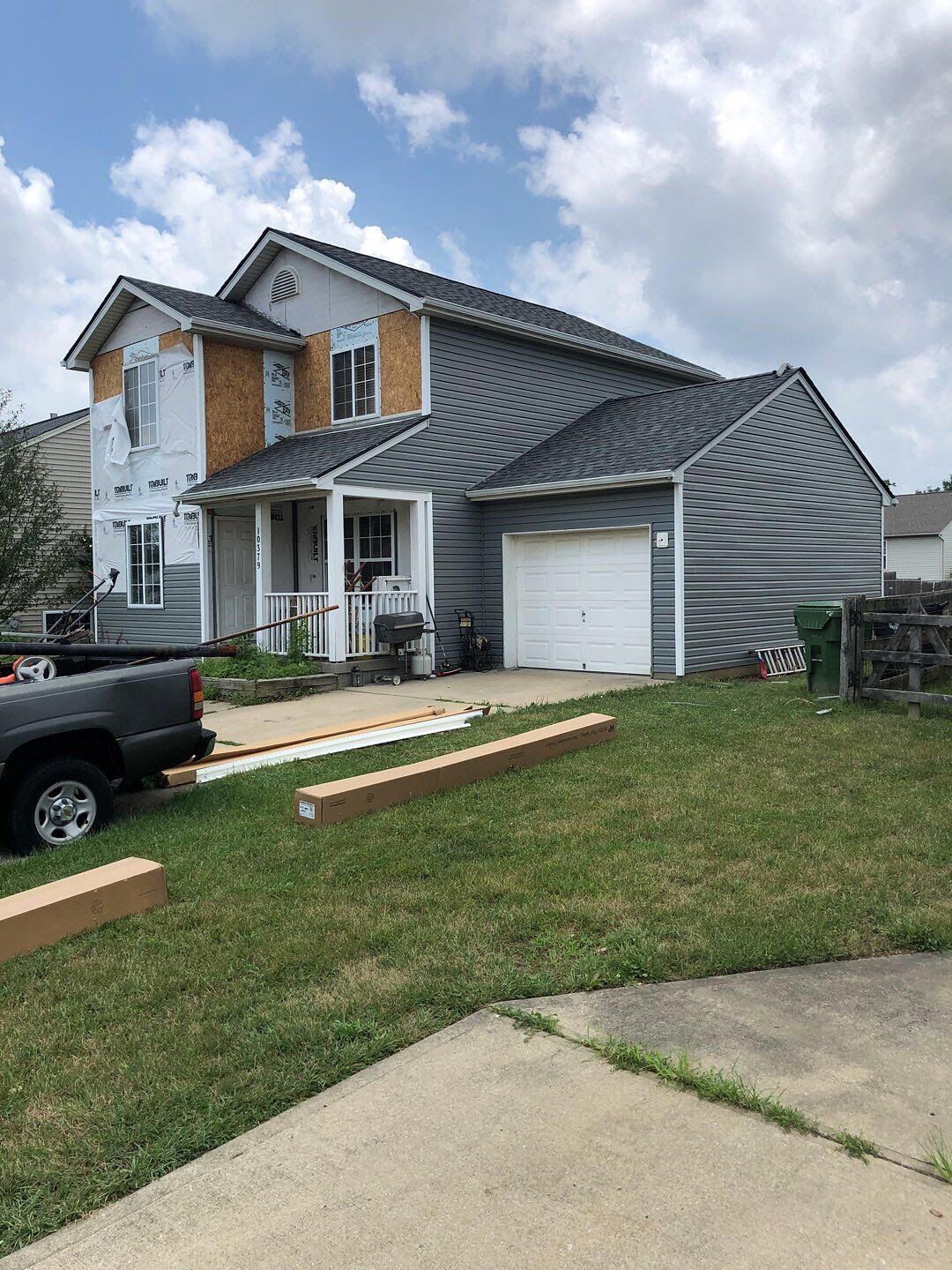 Two-story gray house with white trim, garage, and lumber on the front lawn under a cloudy sky