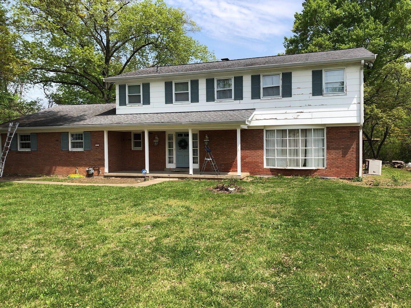 Two-story brick house with white siding, front porch, and large green lawn under trees