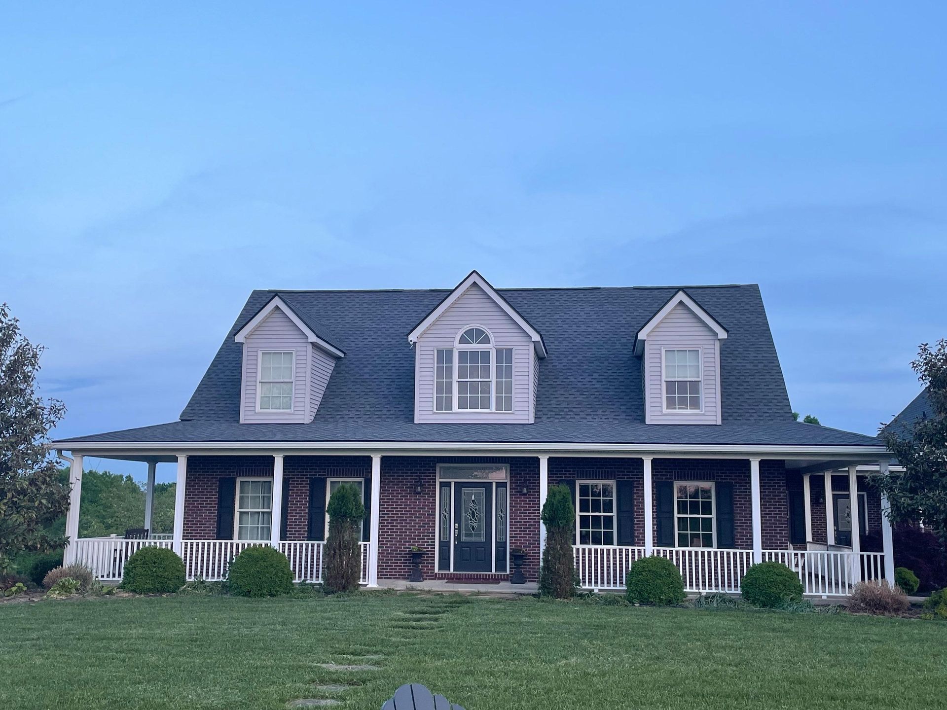 Two-story brick house with white porch, dark roof, and dormer windows under a blue evening sky