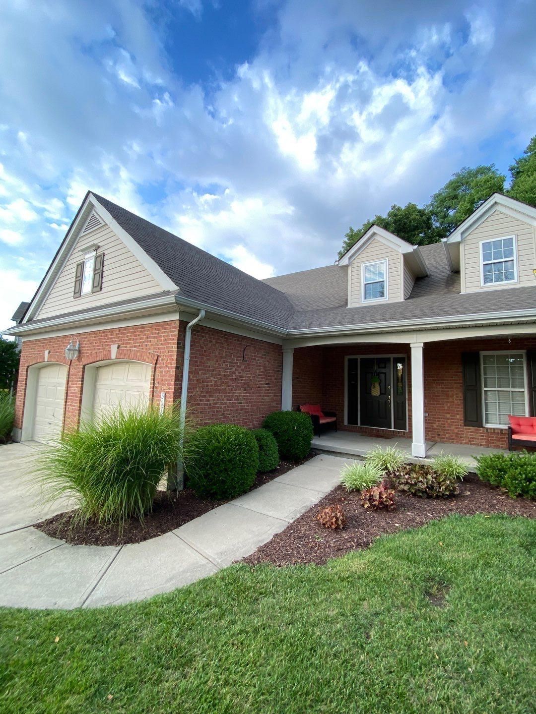 Brick suburban house with red shutters, front porch, and landscaped lawn under a cloudy blue sky