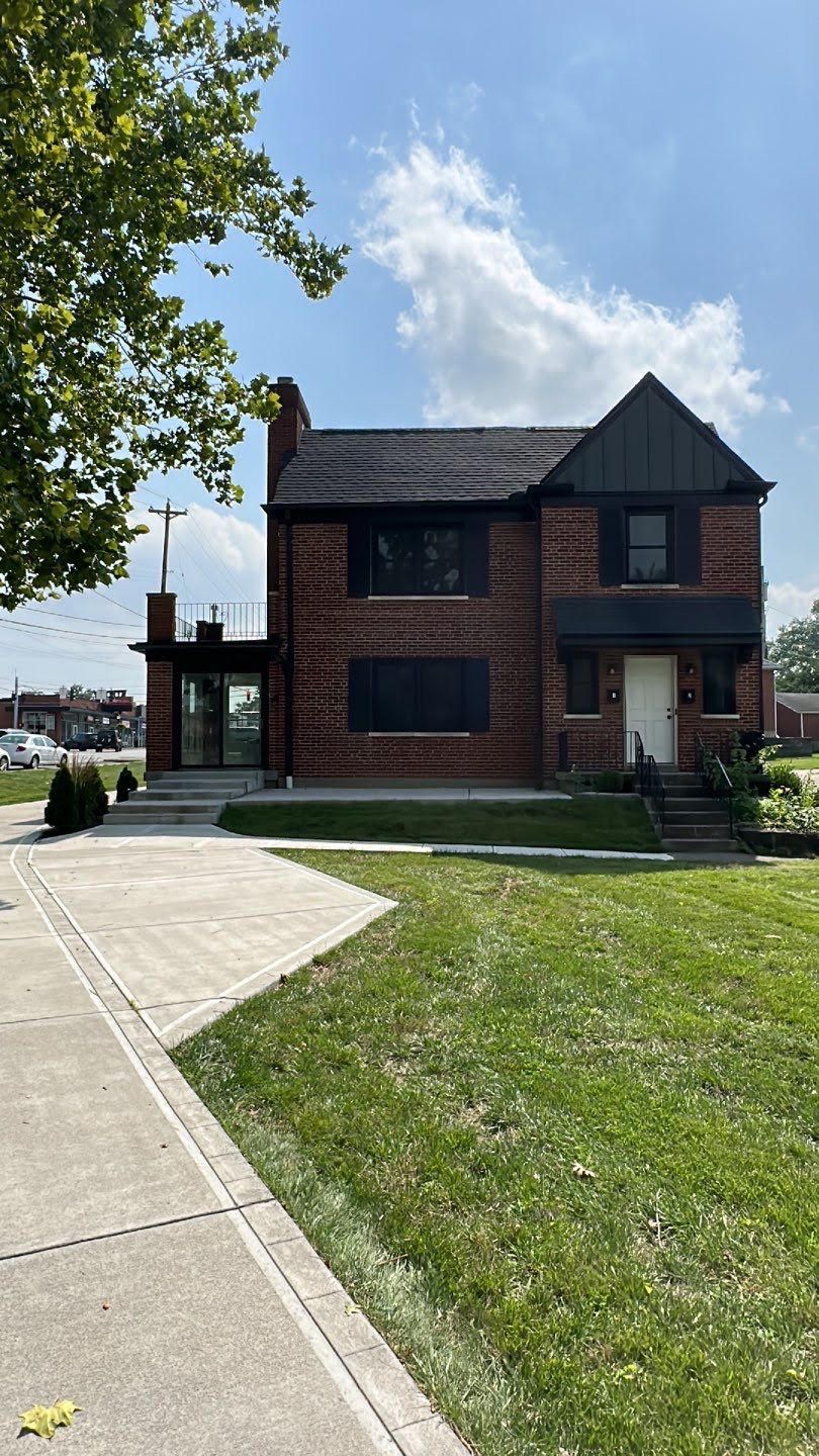 Brick house with red shutters beside a sidewalk and green lawn under a blue sky.