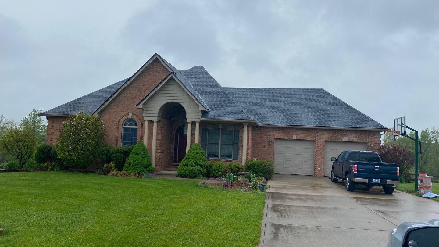 Brick suburban house with two-car garage, green lawn, and a black SUV in the driveway on a cloudy day