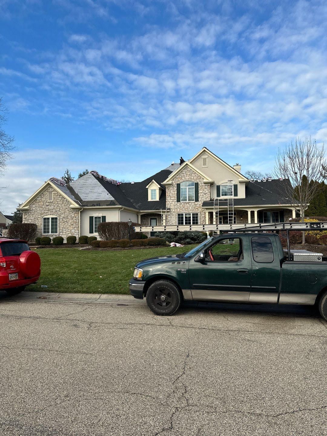 Suburban house with stone facade and parked red and green vehicles under a blue, cloudy sky