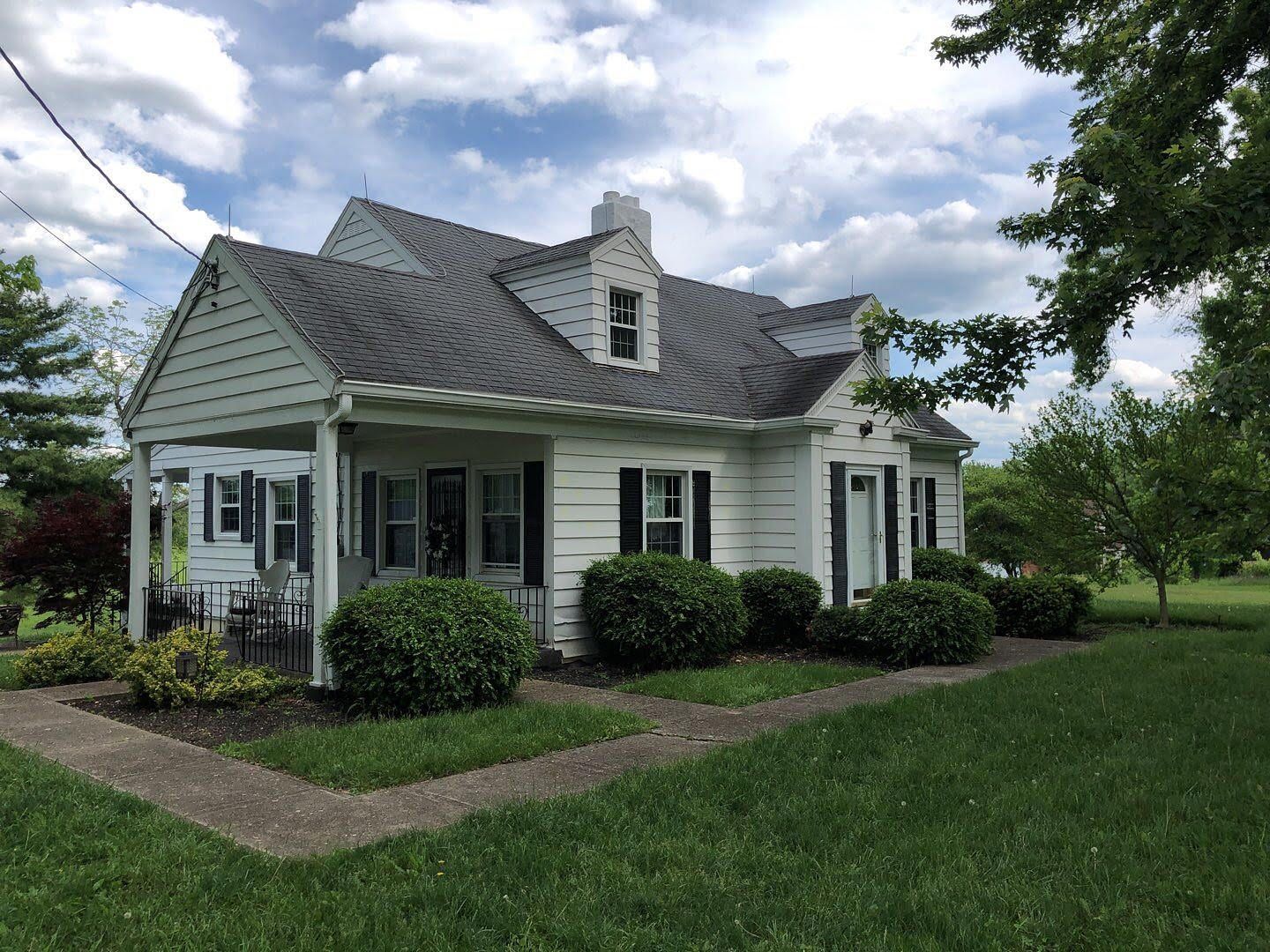 Small white house with green shutters and a gray roof, surrounded by grass and shrubs under a cloudy sky