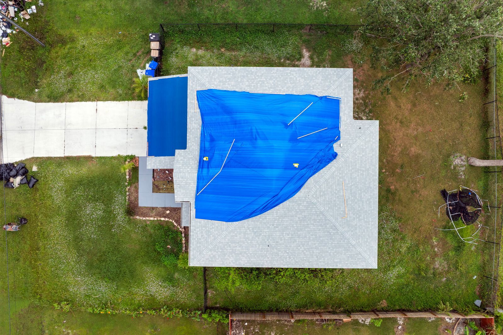 Aerial view of a house roof covered with a blue tarp, surrounded by green lawn and a concrete walkway.