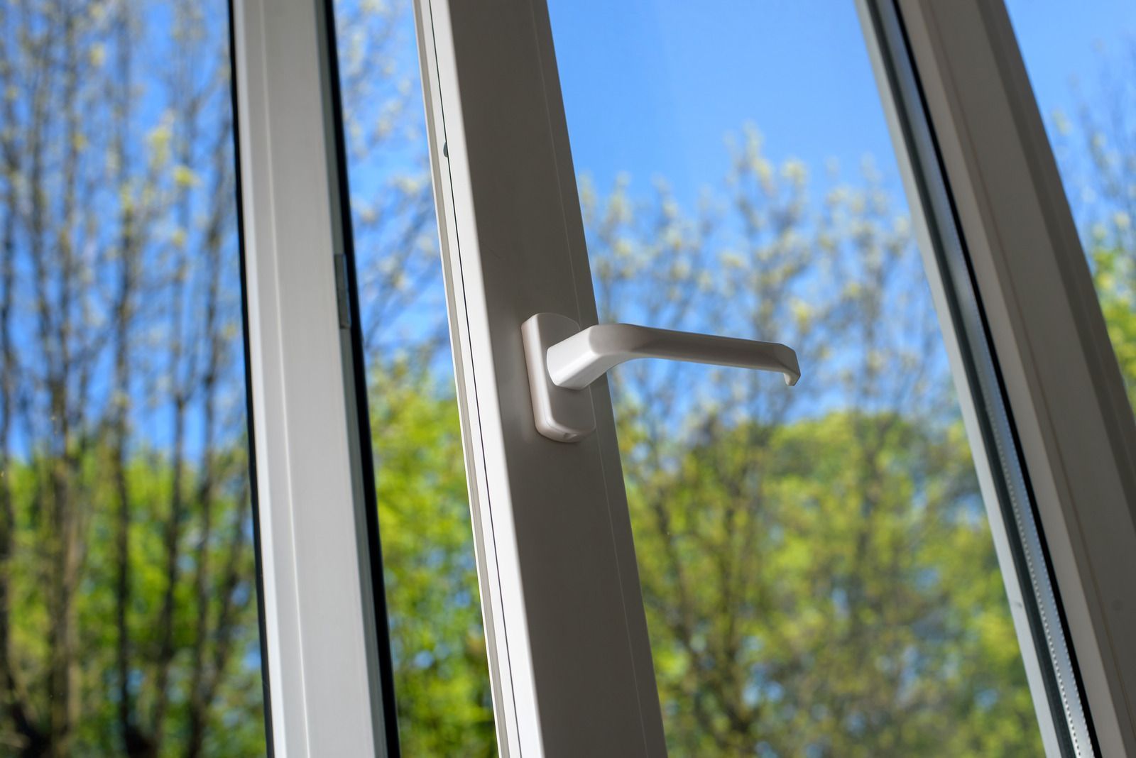 White window handle on a sunlit open window with blue sky and bare trees outside