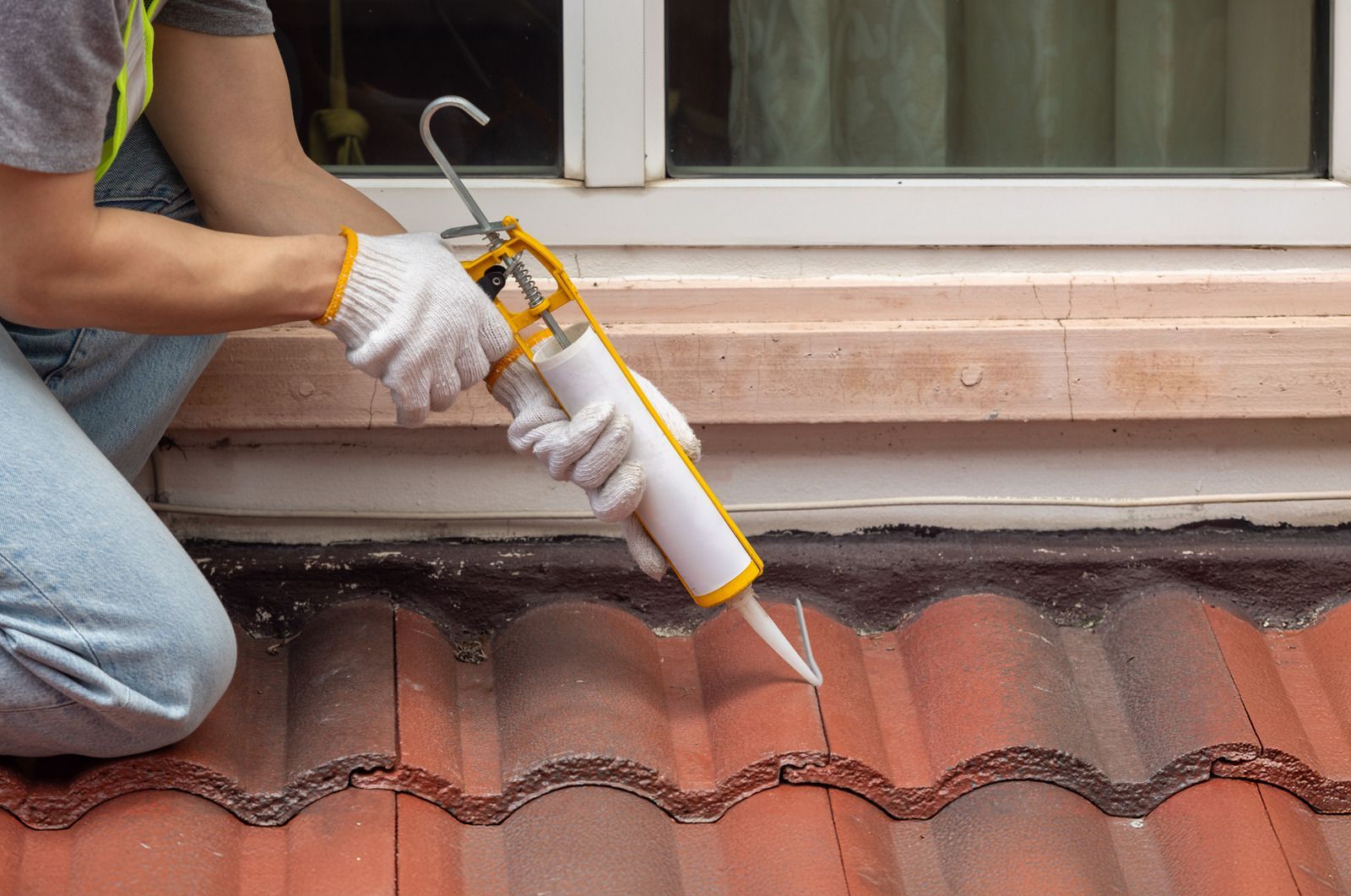 Gloved worker applies sealant to a tiled roof beside a window