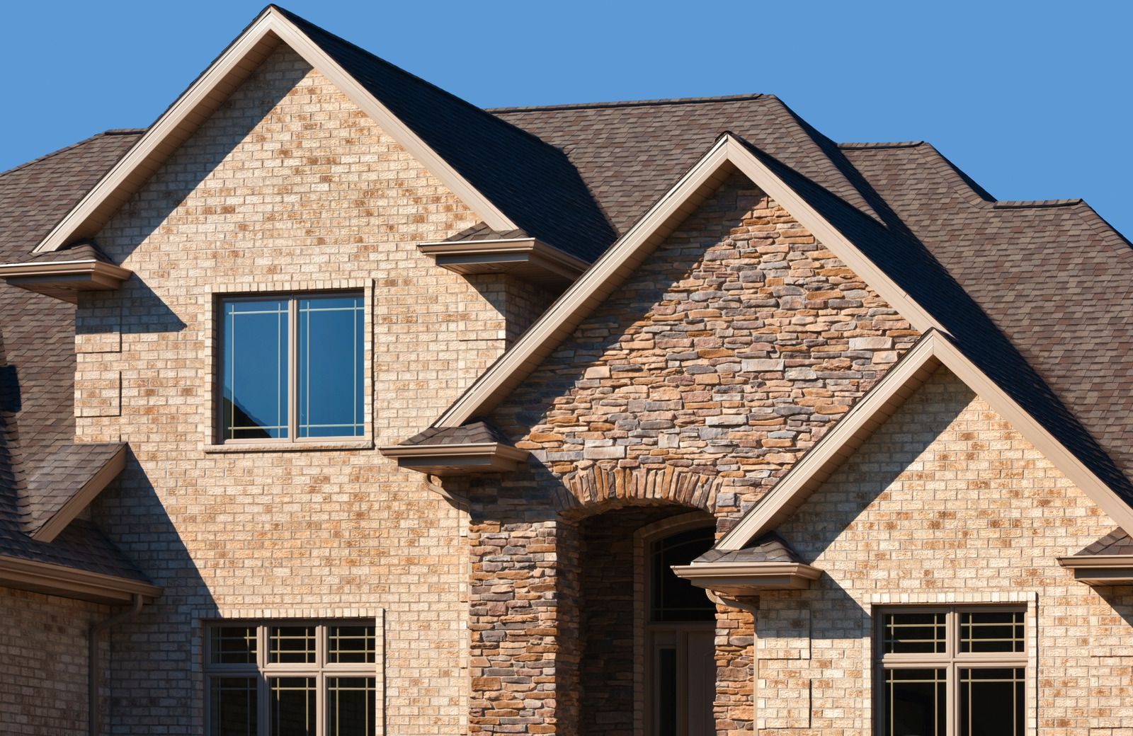 Stone suburban house facade with blue-trimmed windows and multiple gabled roofs under a clear sky