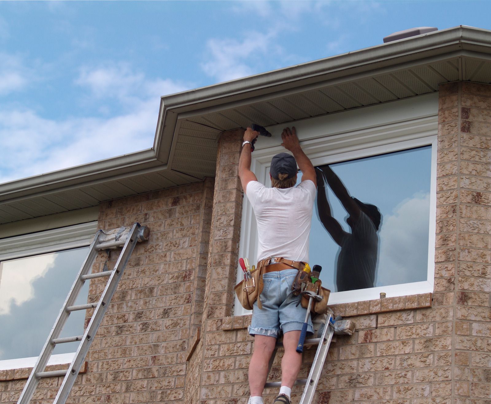 Worker installing a window shutter on a brick house, with ladders leaning nearby.
