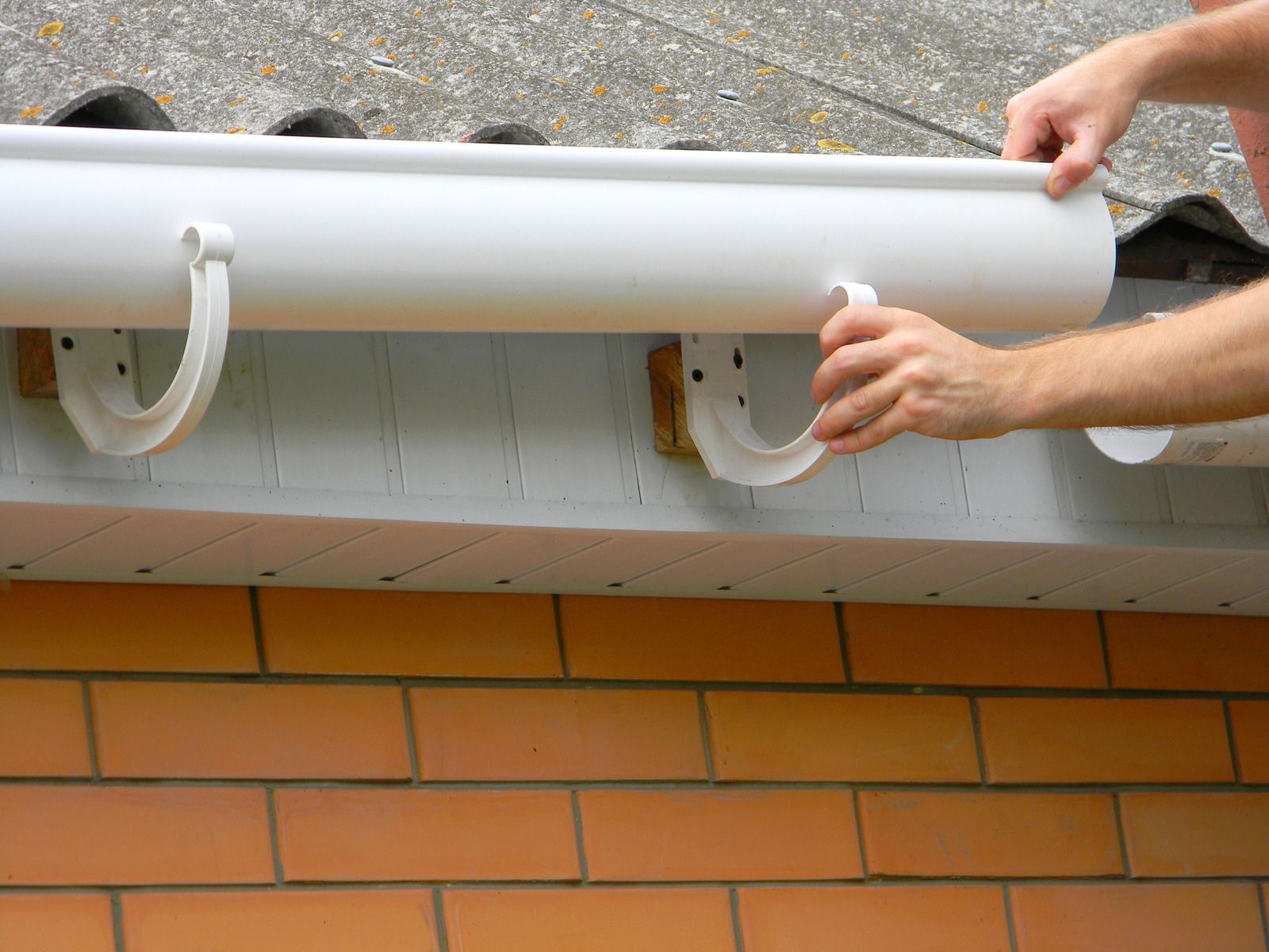 Hands attaching a white gutter or awning to a brick wall under a shingled roof.
