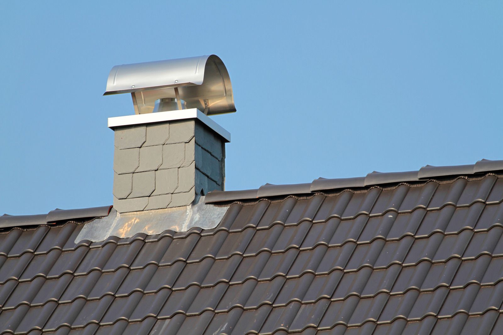Metal chimney vent on a roof with brown shingles against a blue sky