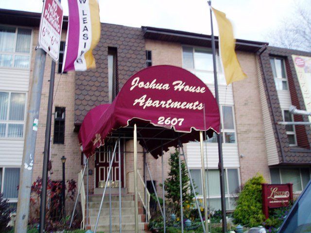 Joshua House Apartments building entrance with burgundy awning, flagpoles, and leasing sign