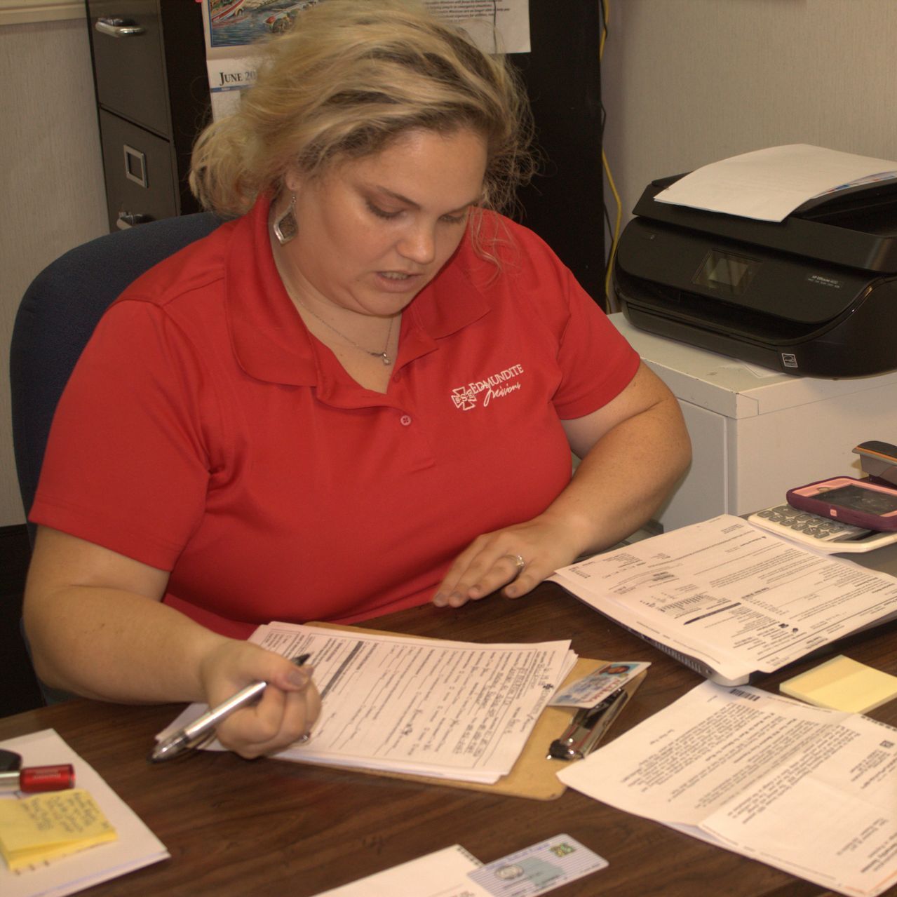 A woman in a red shirt sits at a desk in front of an hp printer