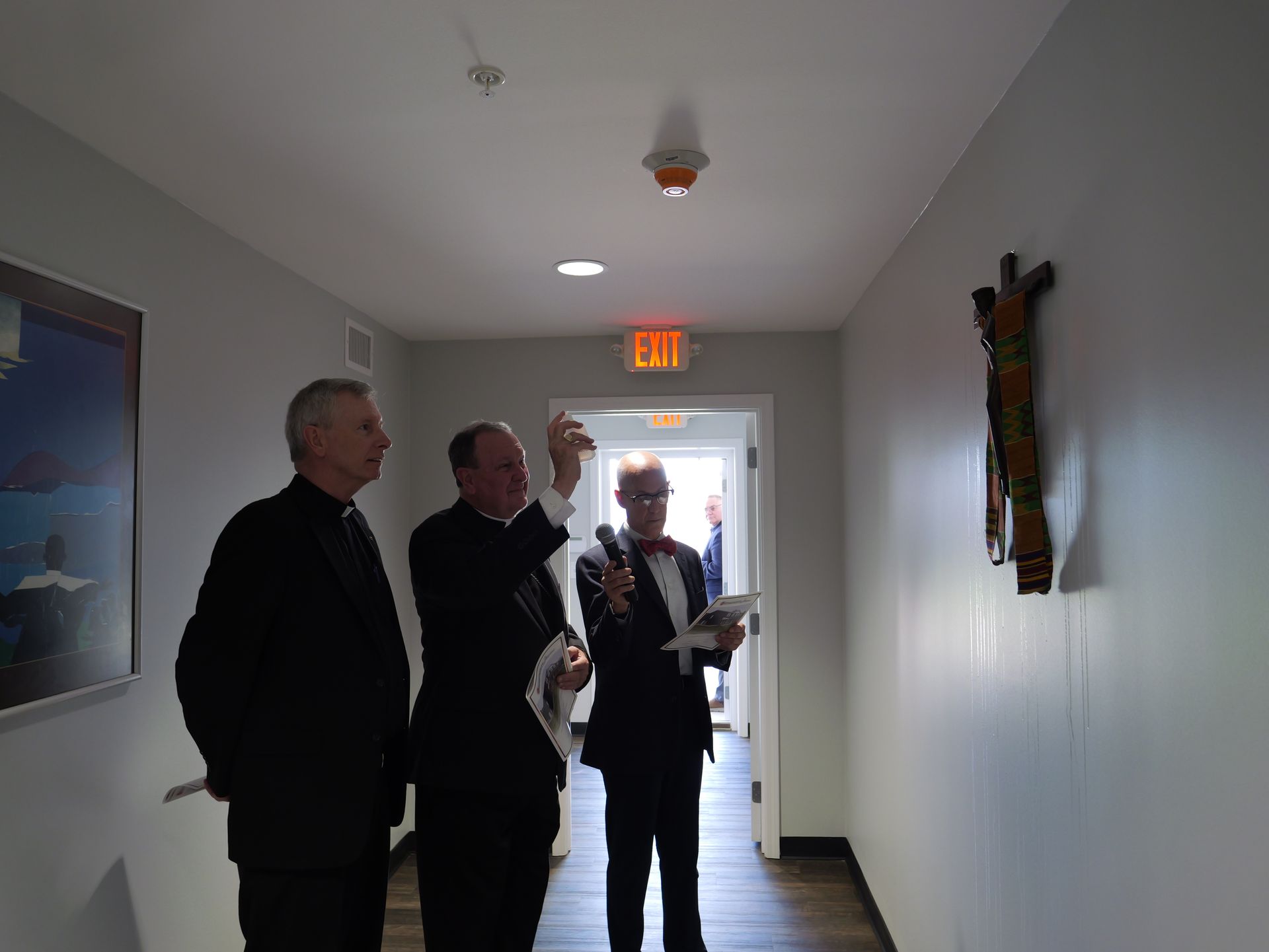 Three men are standing in a hallway looking at a painting on the wall.