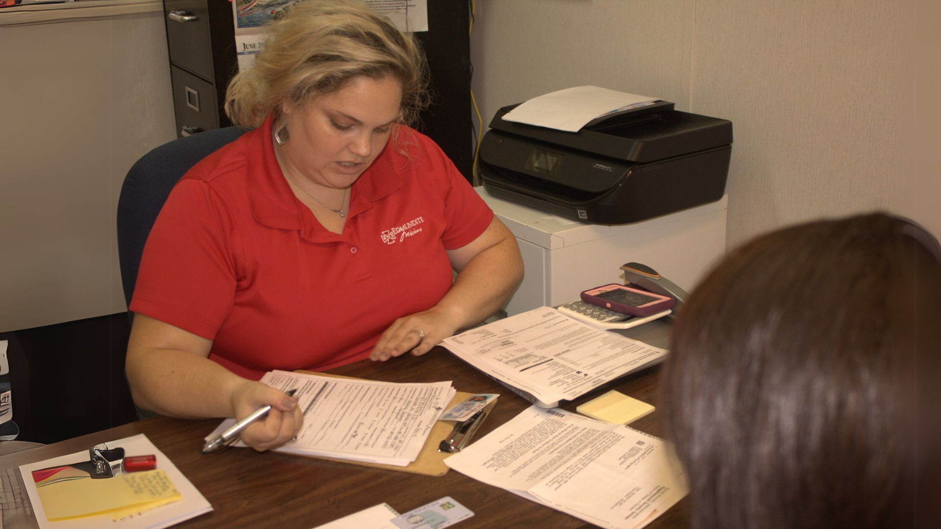 A woman in a red shirt is sitting at a desk with papers