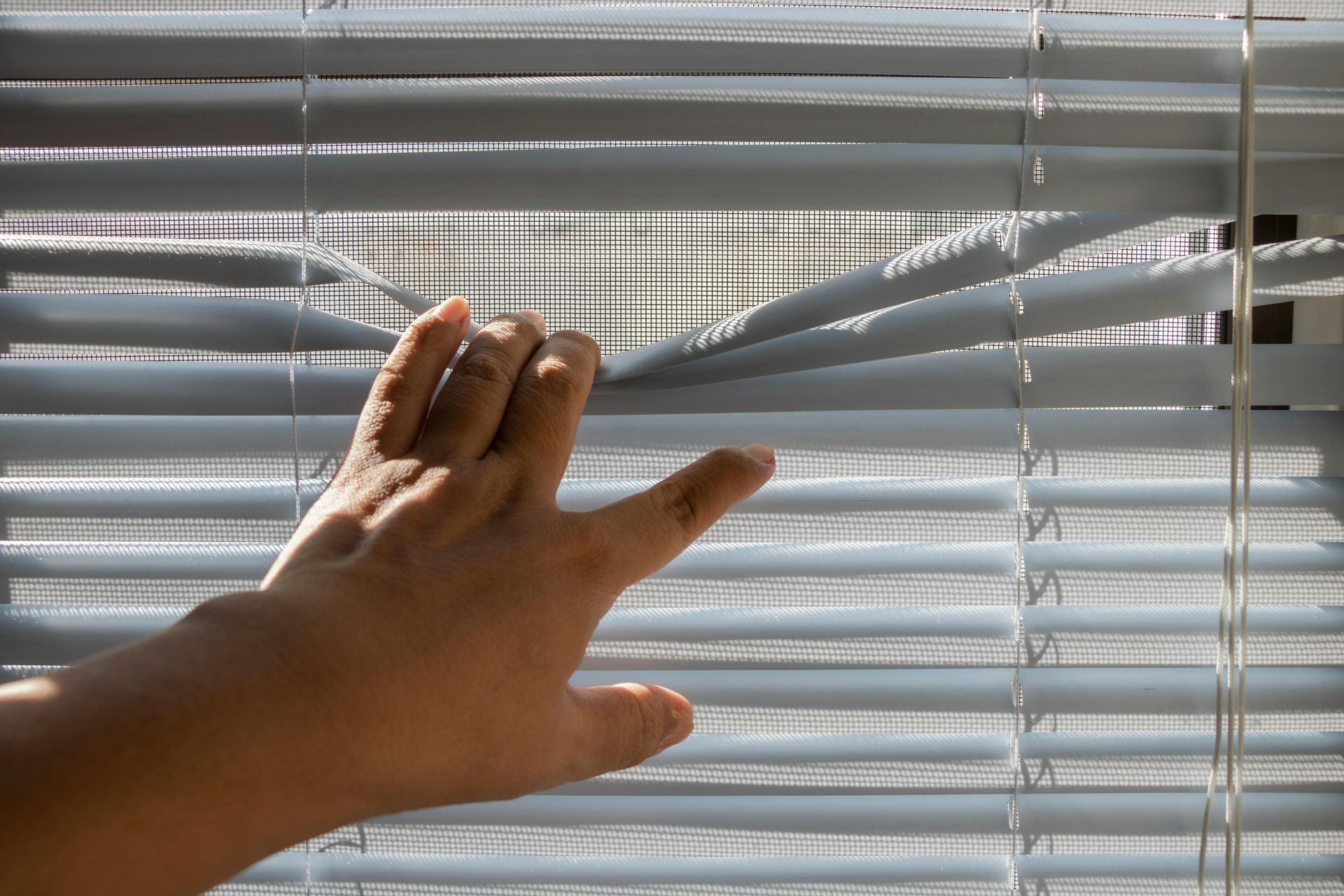 Hand parting closed blinds to peek outside.